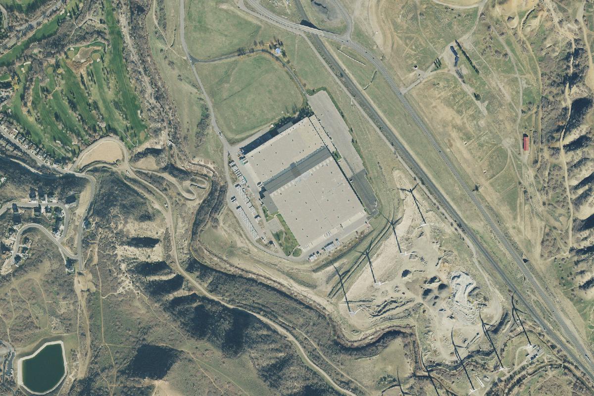 Aerial view of the parcel at 4000 East Highway 6 in Spanish Fork: a large warehouse with a paved yard beside U.S. Route 6, a gravel pit and wind turbines to the south, homes and a golf course to the west.
